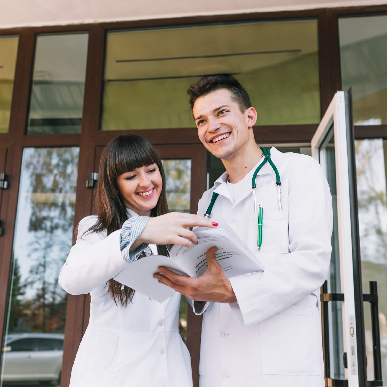 Two smiling medical students in white coats, standing outside a university building, reviewing study materials together. The male student wears a stethoscope, and both appear engaged in discussion, reflecting the collaborative and dynamic learning environment typical of studying medicine in UK.